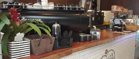 A coffee shop counter with a coffee machine, a menu board, and a brick wall with a logo.
