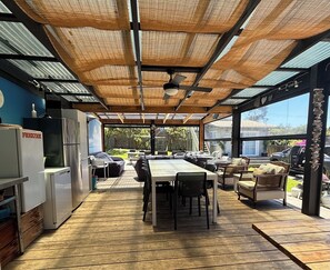 An outdoor dining area with a wooden deck, metal-framed ceiling, and large windows.