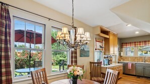 Un comedor con una mesa redonda de madera, seis sillas y una araña. Hay ventanas grandes con cortinas, vistas al exterior y una zona de cocina con un fregadero y armarios.