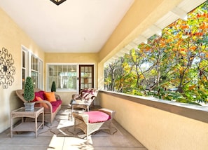 A sunlit balcony with wicker furniture, a small potted tree, and a view of autumn foliage.