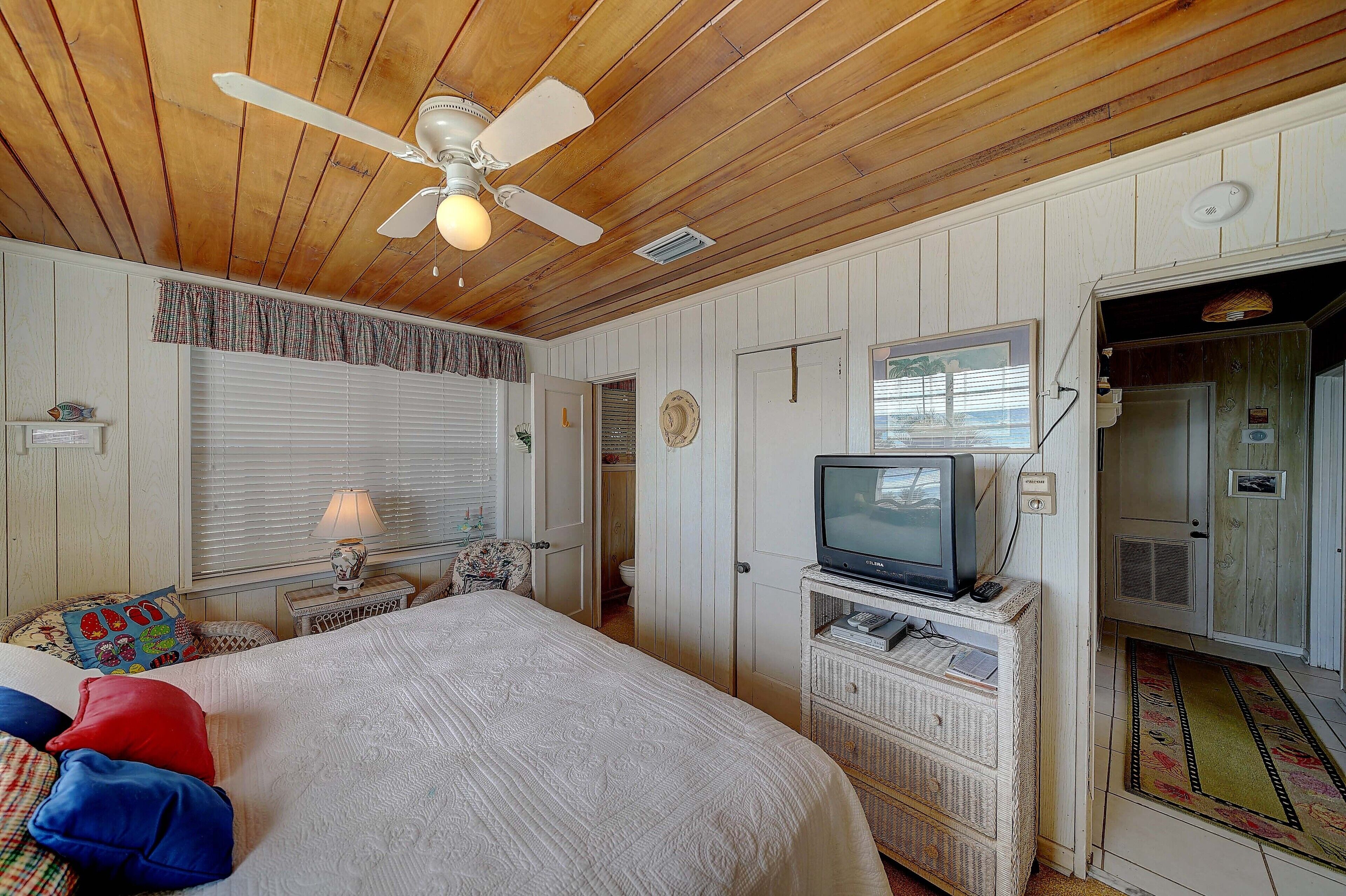 A bedroom with a bed, a ceiling fan, a television on a stand, and a door leading to another room.