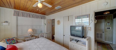 A bedroom with a bed, a ceiling fan, a television on a stand, and a door leading to another room.