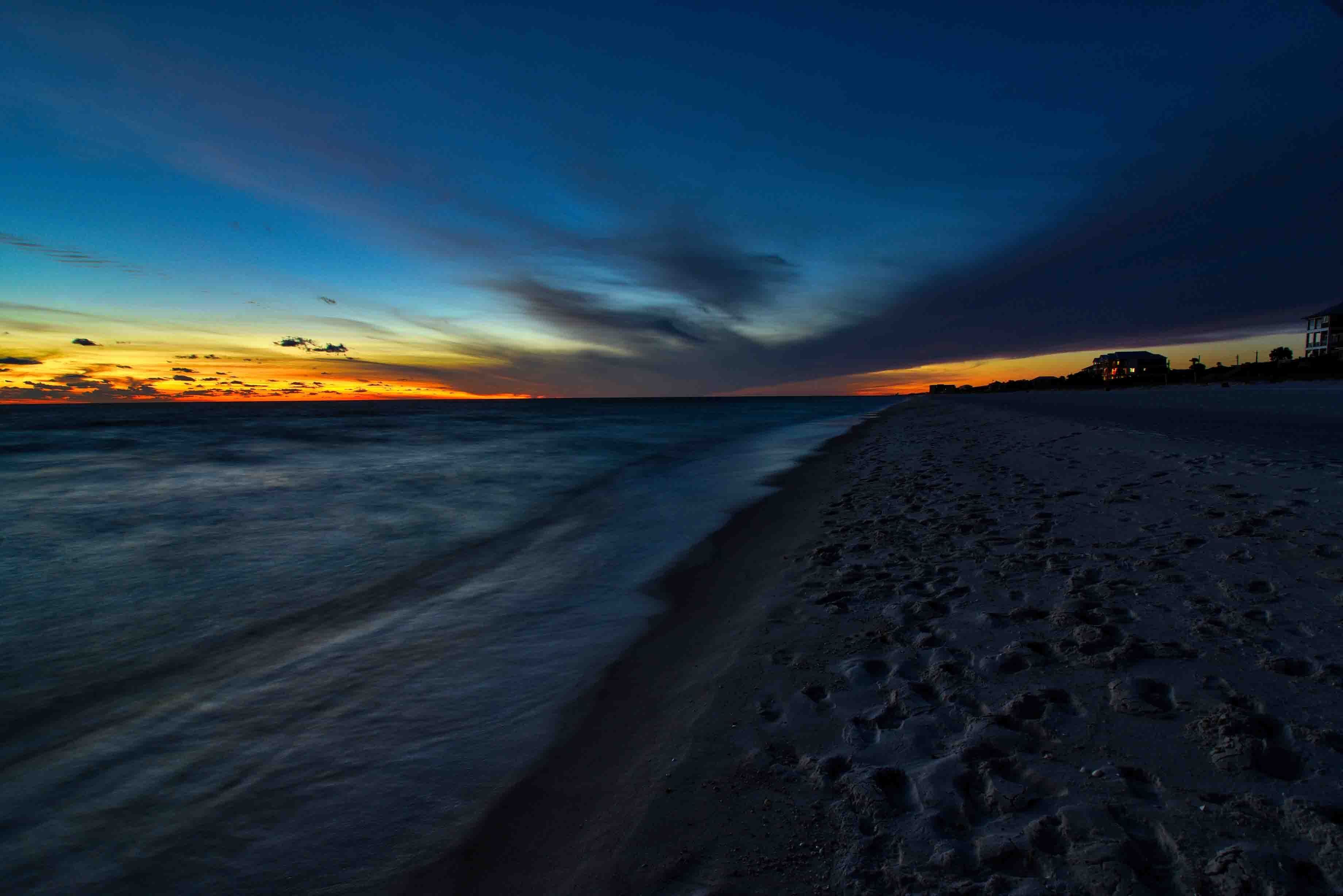 A beach with footprints leading to the water, a sunset in the background, and a building on the right.