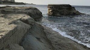 A coastal landscape with rocky formations and a distant view of buildings. - Cosy Air-conditioned Studio Marsalforn Beach (Marsalforn, Żebbuġ Gozo)