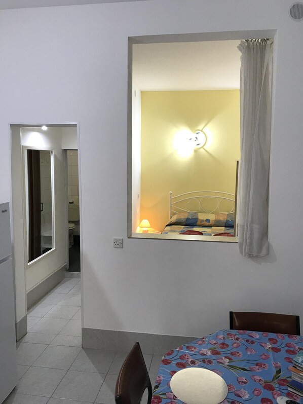 A small room with a bed, a window with curtains, a table with a floral tablecloth, and a chair.