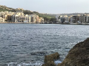 A coastal cityscape with buildings along the waterfront, rocky shoreline, and a hilly background.