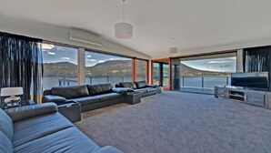 Ein geräumiges Wohnzimmer mit einer großen Sofagarnitur, einem Flachbildfernseher und Blick auf ein Gewässer und Berge.