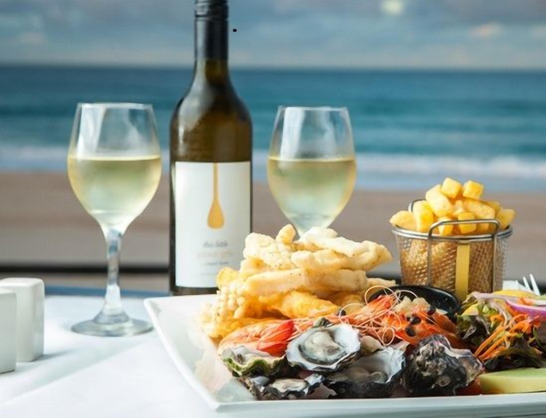 A table with seafood dishes, a bottle of wine, and glasses of white wine, set against a beach view.