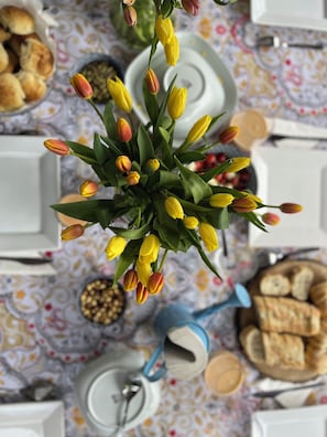 A table with a floral tablecloth, a vase of yellow tulips, and various food items including bread and a bowl of nuts.