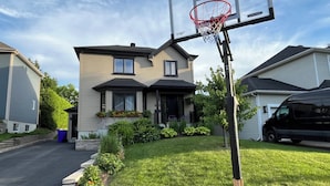 A residential area with a basketball hoop in the foreground, houses with sloped roofs, and a driveway leading to a garage.
