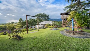 A grassy area with playground equipment, a wooden structure, and a house with a thatched roof. - Khao Kho Valley Hotel (Khao Kho)