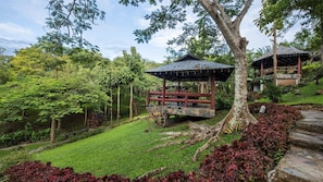 A gazebo with a stone base and wooden railings, surrounded by lush greenery and a small pond.