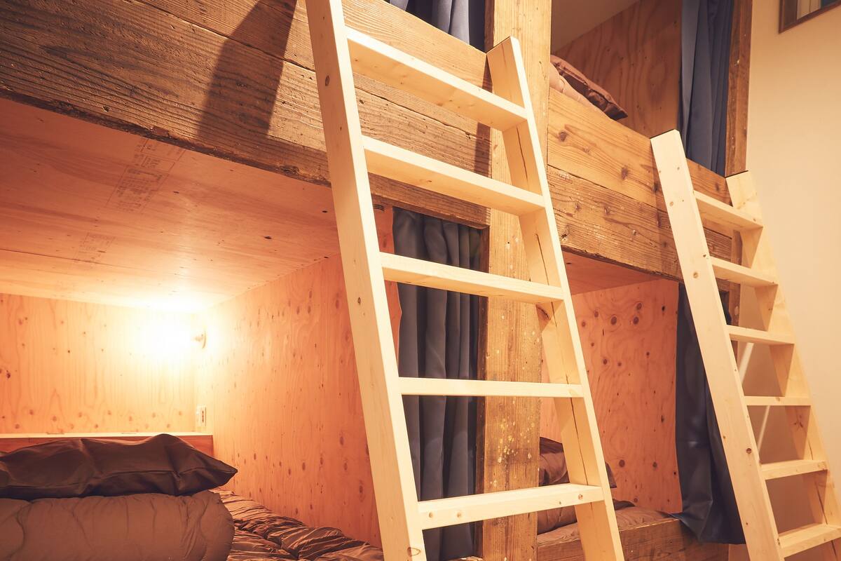 Una cama litera de madera con escalera, un pequeño cofre de madera y una ventana con cortinas.