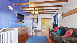 Un salon doté d’un plancher en bois, d’un tapis bleu, d’un canapé en cuir marron agrémenté de coussins colorés, d’une rambarde blanche, d’un téléviseur fixé au mur et d’un escalier en bois.
