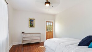 Une chambre avec un plancher en bois, un lit blanc avec des oreillers bleus, une commode, une porte et un ventilateur de plafond.