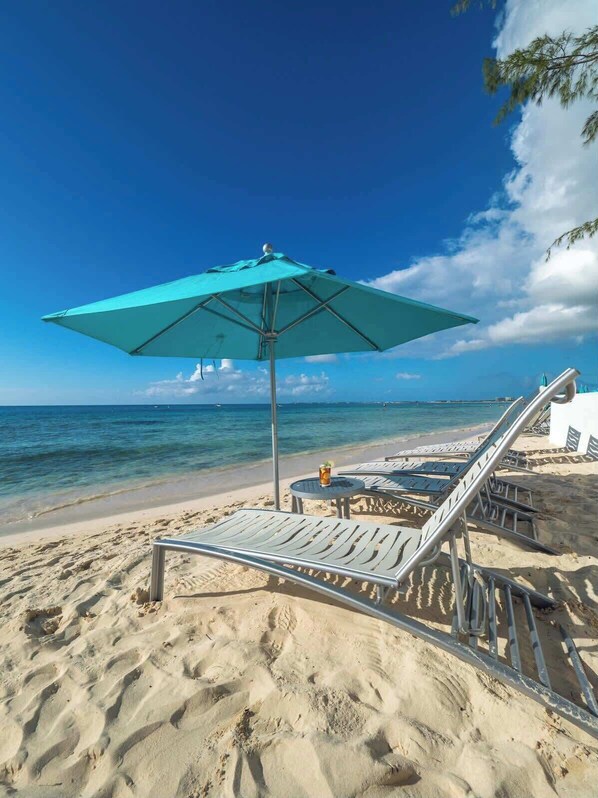 A beach with a turquoise umbrella, lounge chairs, and a view of the ocean.