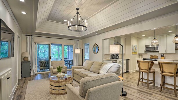 A spacious living room with a large beige sectional sofa, a coffee table, and a dining area with bar stools. The kitchen features white cabinets, a microwave, and pendant lighting. - Walk to Beach! Remodeled Cottage on Sparrow Pond (Kiawah Island)