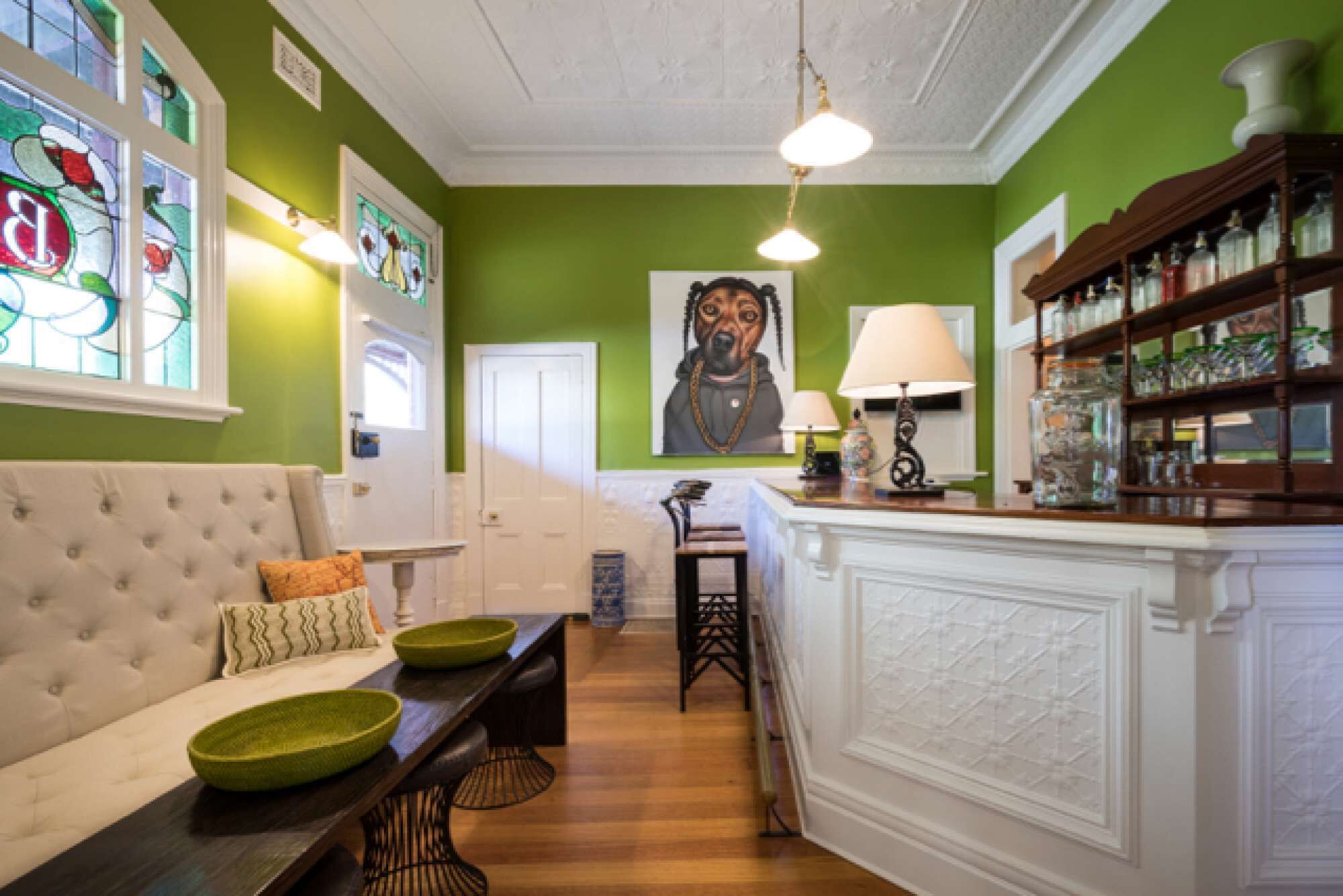 Una habitación con pared verde, un sofá blanco, una mesa de comedor de madera con cuencos verdes, un armario blanco con frascos de vidrio y un cuadro de un perro.