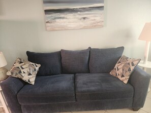 Ein dunkelblaues Sofa mit gemusterten Kissen, ein Gemälde mit Strandmotiv und eine Lampe auf einem Beistelltisch.