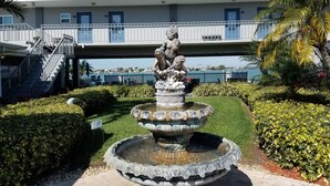 Ein mehrstufiger Steinfbrunnen mit einer Statue in der Mitte, umgeben von gepflegten Hecken und einem gepflasterten Weg.
