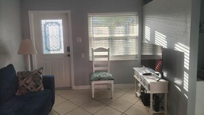 Ein Wohnzimmer mit Sofa, Sessel, Schreibtisch, Fernseher und einem Fenster mit Jalousien.