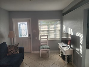 Sala de estar com sofá, poltrona, escrivaninha, televisão e janela com persianas.