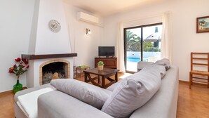 A living room with a fireplace, a television, a sofa, a table with a plant, and a view of a pool through a glass door. - Casa Prensa - Private pool, 5 minute drive to Carvoeiro & beach (Lagoa)