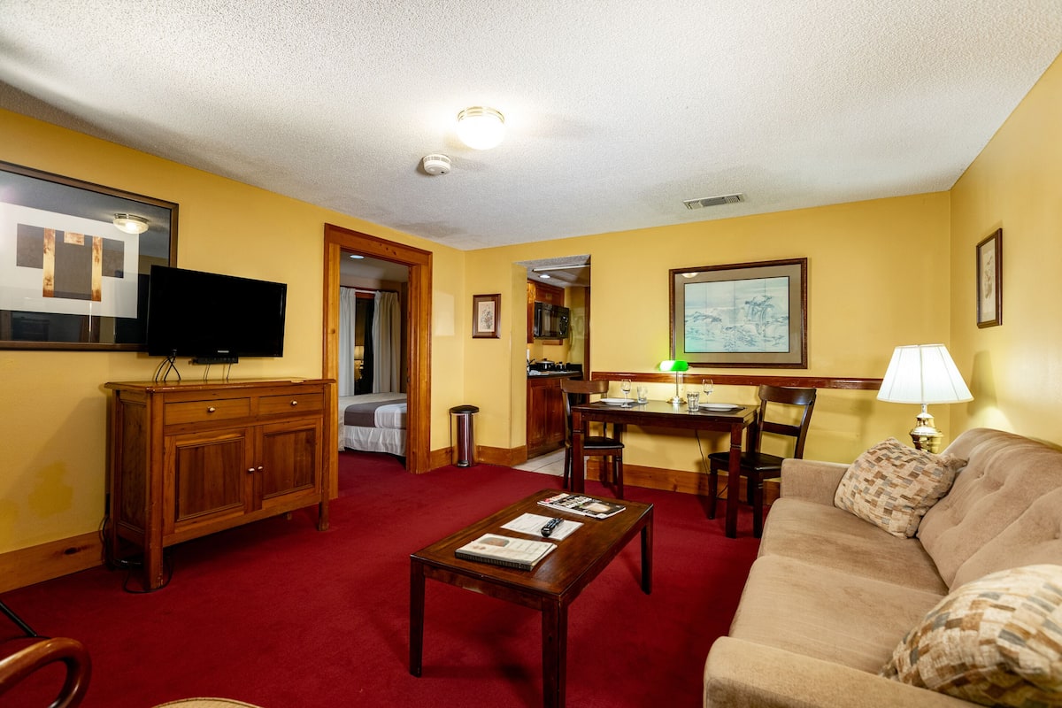 Habitación de hotel con alfombra roja, muebles de madera, televisor, zona de comedor y un cuadro en la pared.