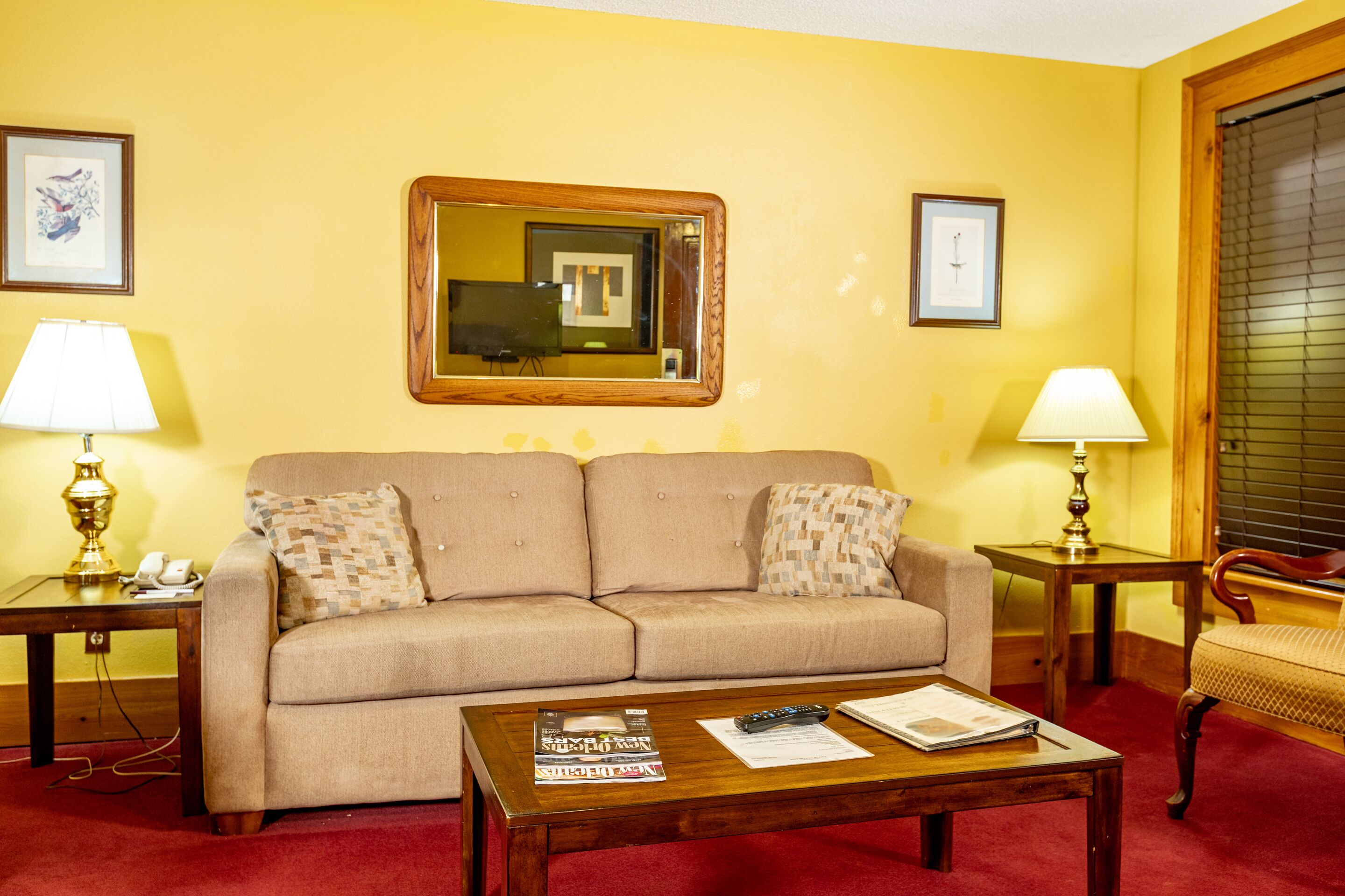 A living room with a beige sofa, two side tables with lamps, a wooden coffee table with magazines and a remote control, and a large mirror on the wall.