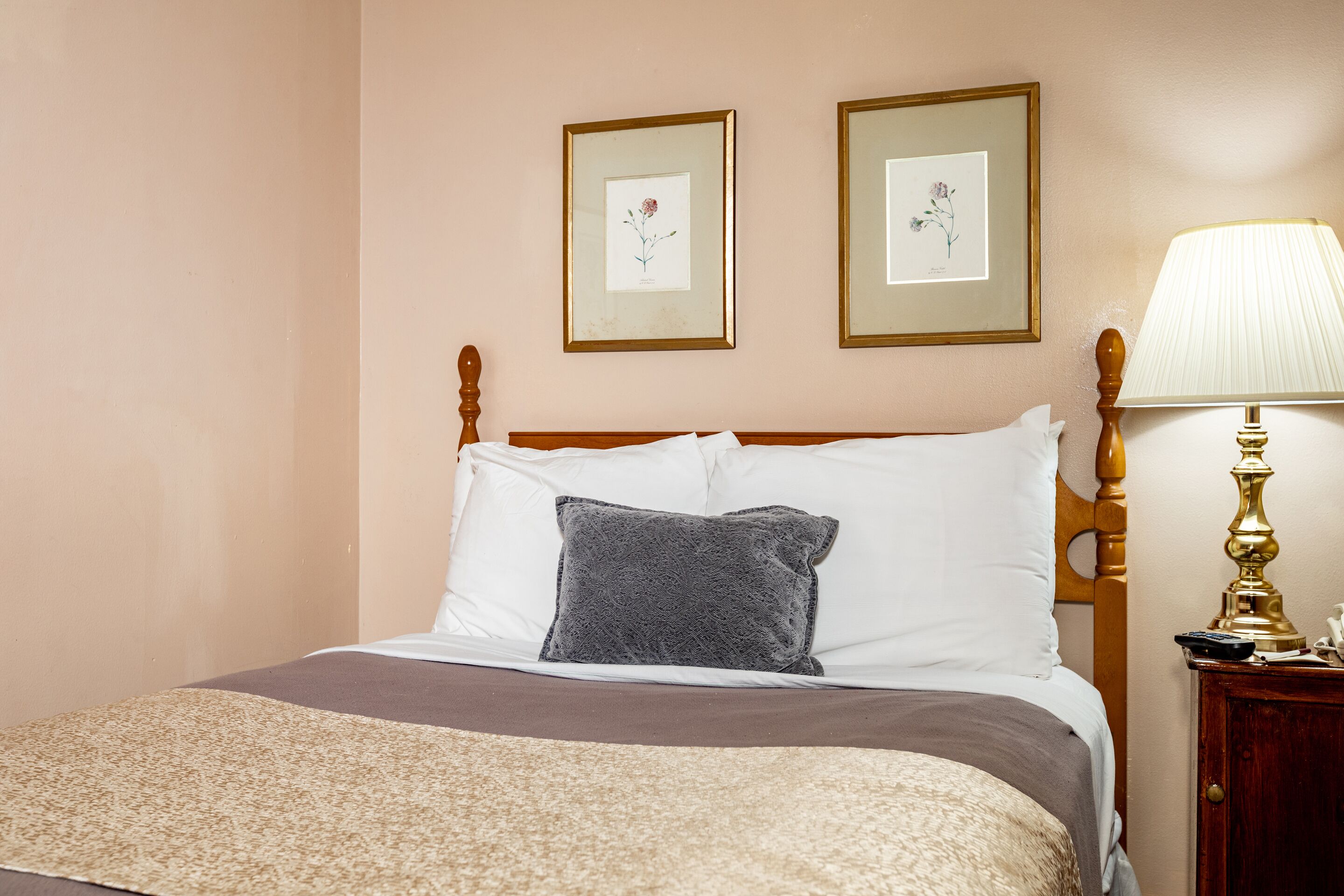 A neatly made bed with a wooden headboard, a gray pillow, and a beige blanket. There are two framed botanical prints on the wall above the bed.