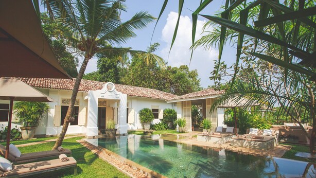 Un espace piscine avec des chaises longues, une piscine carrelée et un bâtiment blanc doté d’un toit également carrelé.