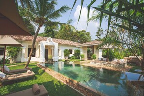 Uma área de piscina com espreguiçadeiras, uma piscina azulejada e um edifício branco com telhado ladrilhado.