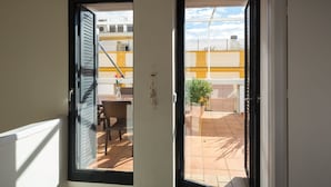 Een geopende deur die leidt naar een balkon met een stoel, een tafel en een potplant.