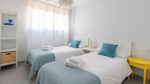Een hotelkamer met twee bedden, een witte kledingkast, een geel nachtkastje en een groot raam met dunne gordijnen.