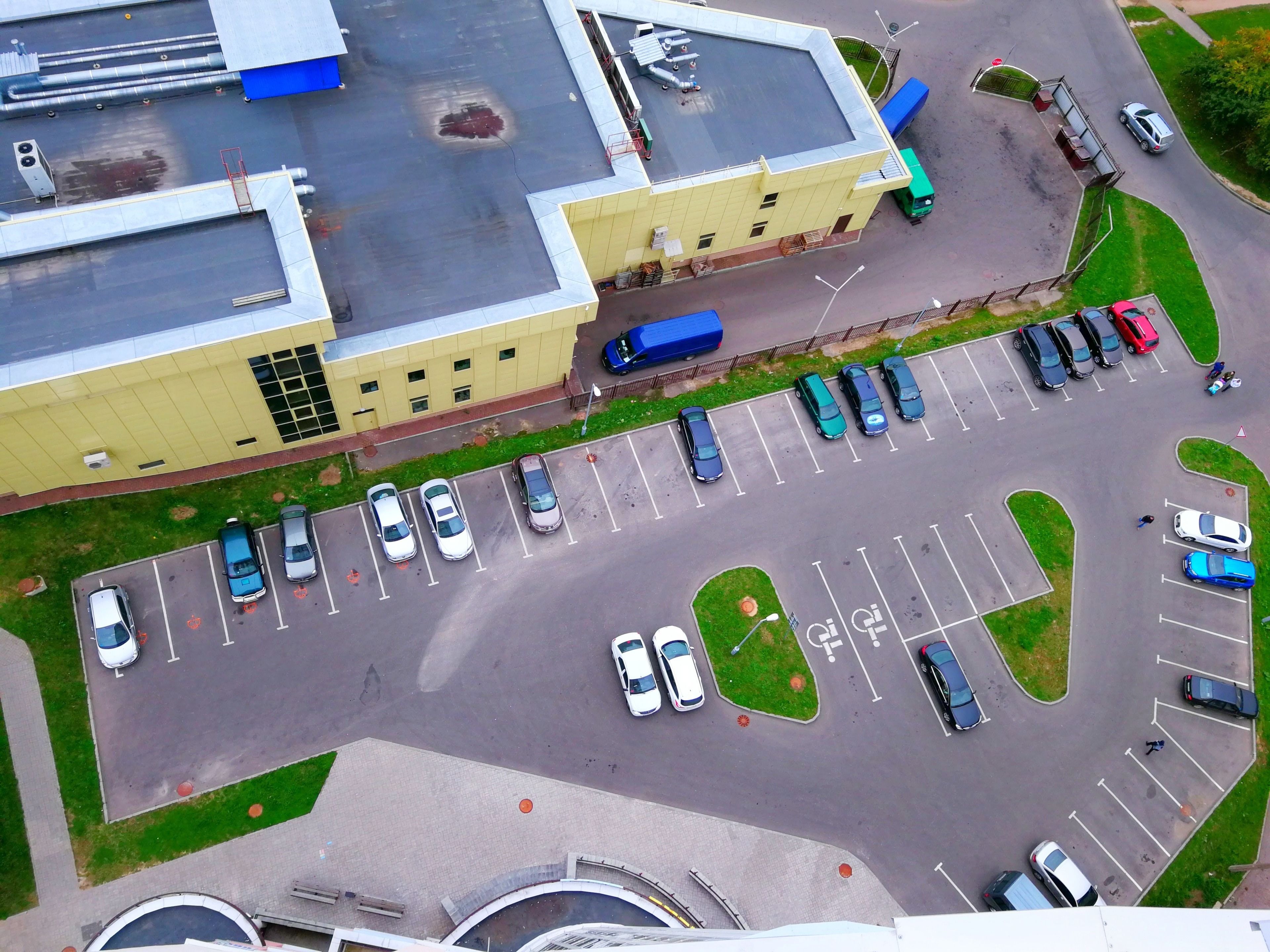 An aerial view of a parking lot with multiple rows of parked cars, a building with a flat roof, and a green area with a small circular patch of grass.
