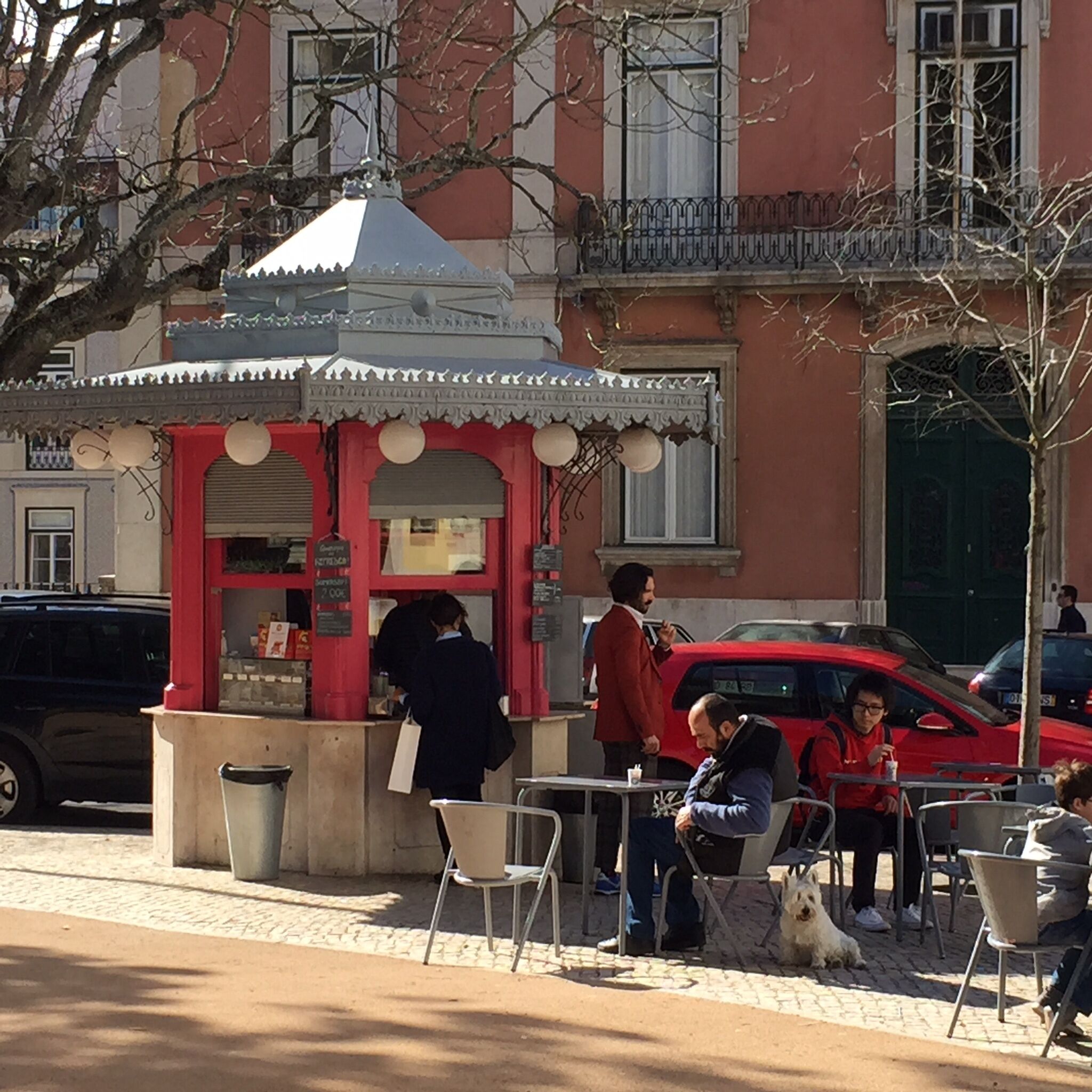 Een café aan de straat met terras, een rood gebouw op de achtergrond en een geparkeerde auto.
