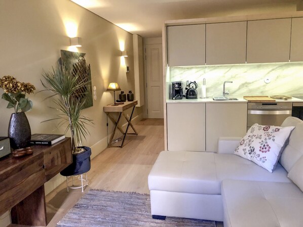 Uma sala de estar moderna com um sofá branco, uma mesa de centro e uma pequena mesa lateral com uma luminária. Há uma área de cozinha com bancada de mármore e uma lava-louças.