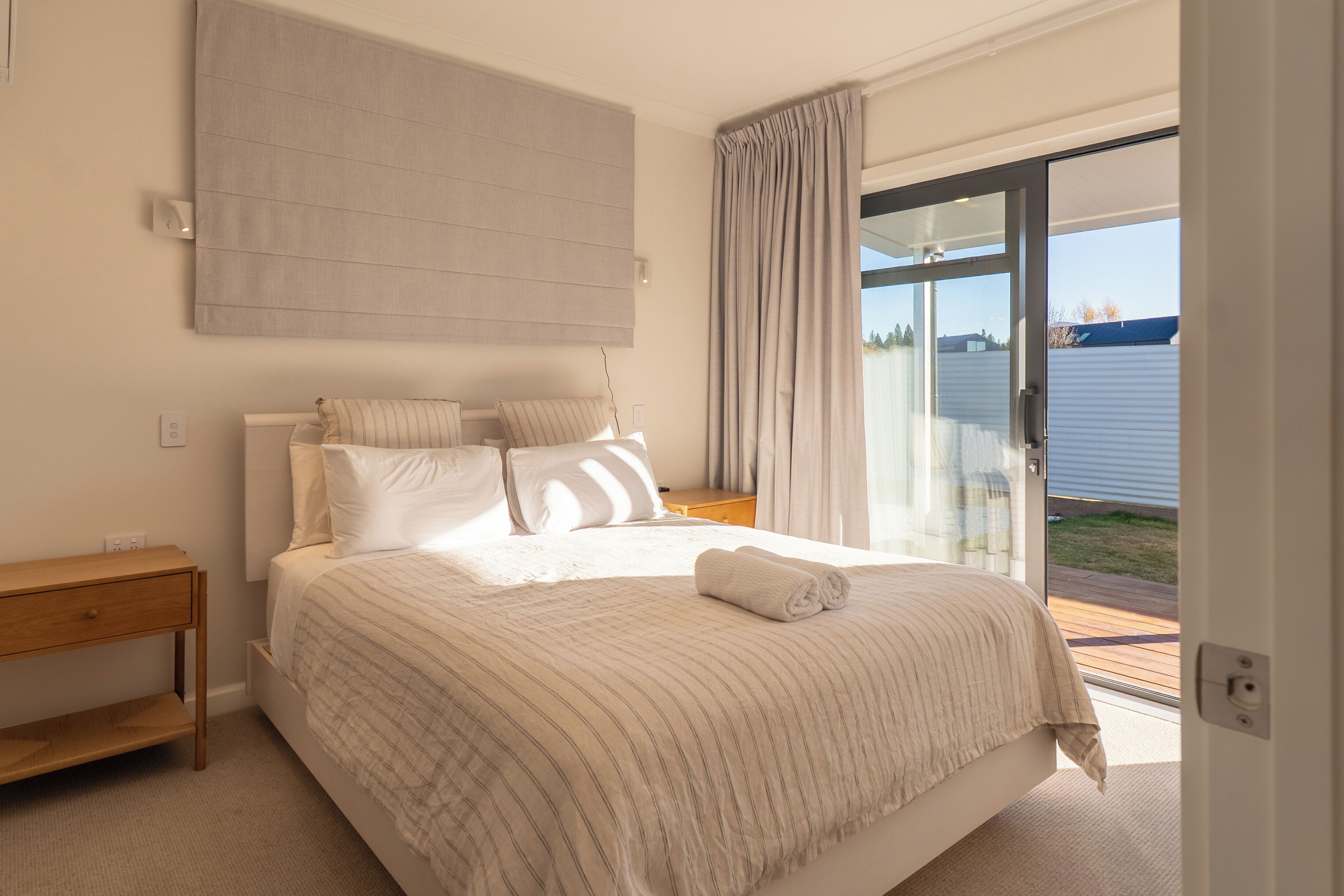 A bedroom with a large bed, a bedside table, a nightstand, and a view of the outdoors through a sliding glass door.