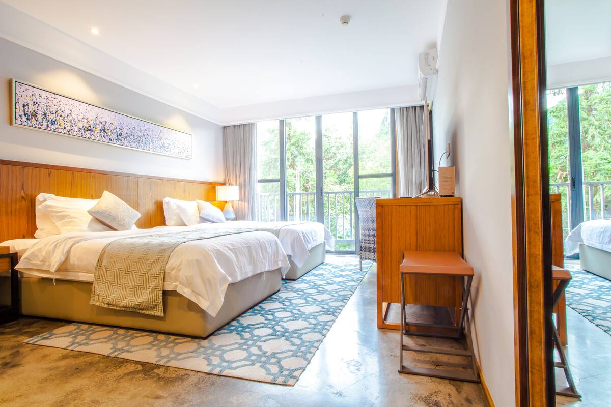 A modern hotel room with a large bed, a desk, and a balcony with a view of greenery.