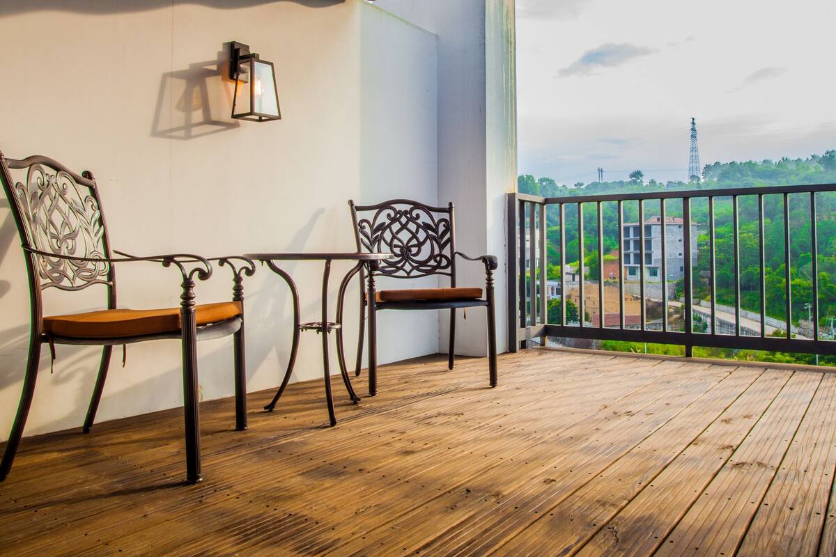 A balcony with wooden flooring, two chairs, a table, and a wall-mounted light fixture.
