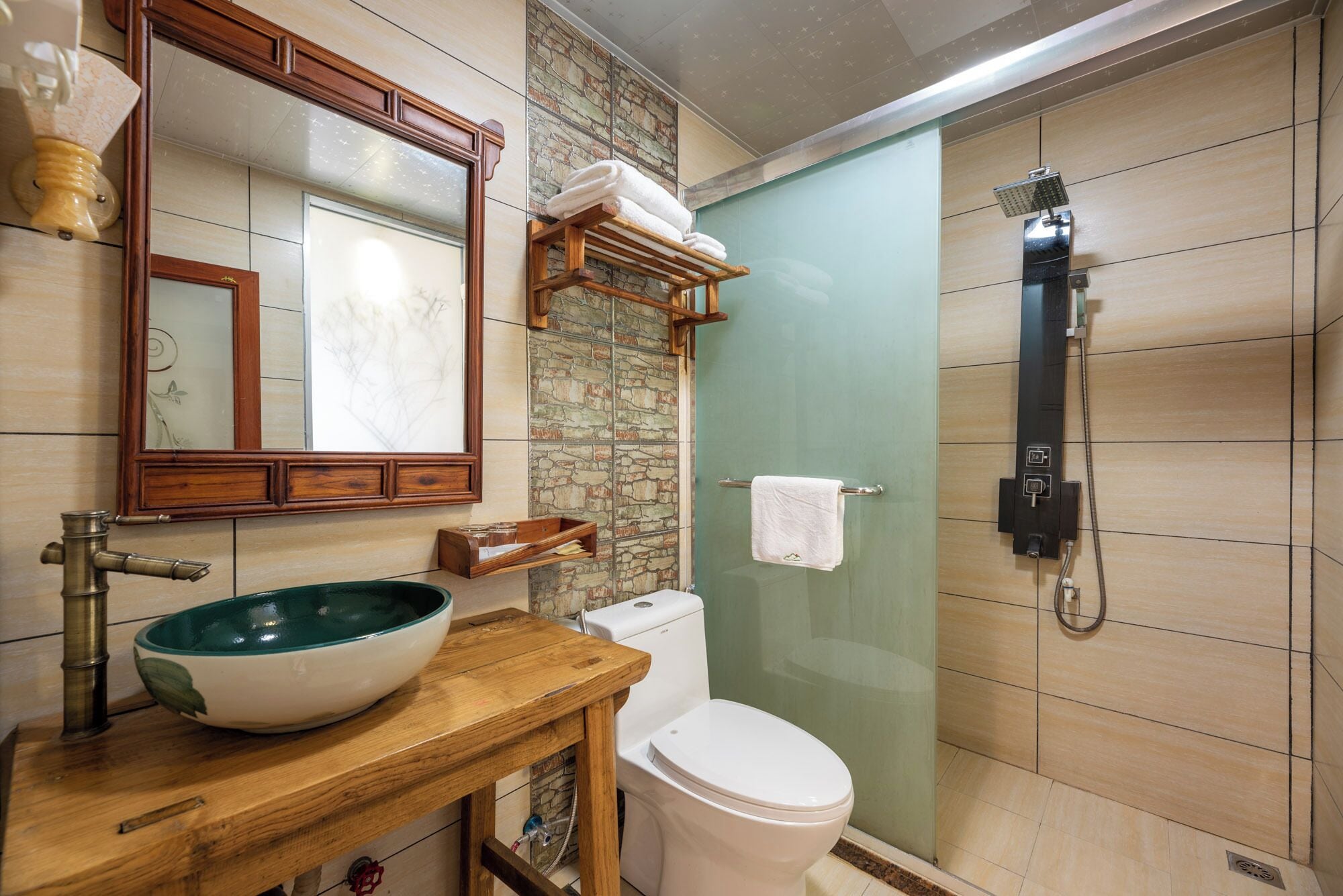 A bathroom with a wooden vanity, a large mirror, a stone accent wall, a toilet, and a glass-enclosed shower.