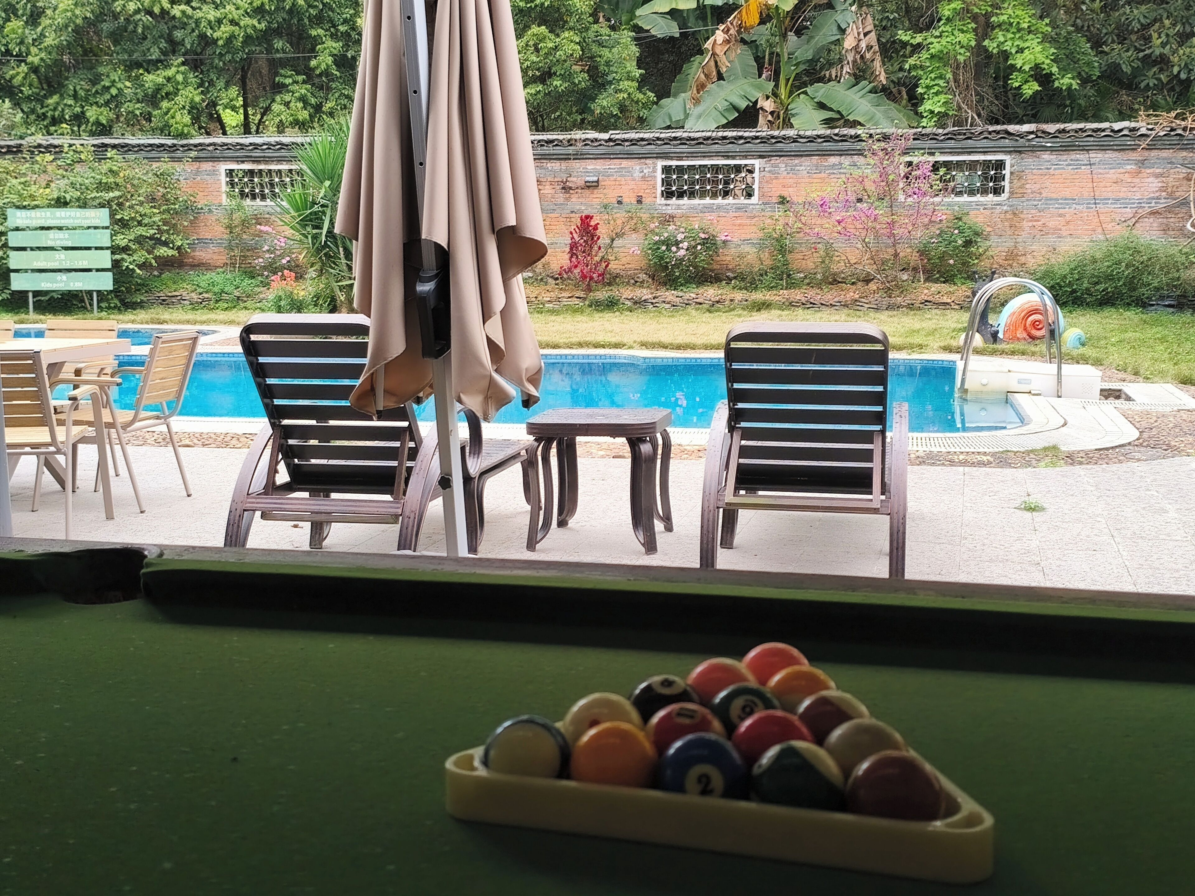 A pool table with billiard balls, a pool table, and two lounge chairs with a pool table in the background.