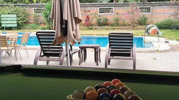 A pool table with billiard balls, a pool table, and two lounge chairs with a pool table in the background.