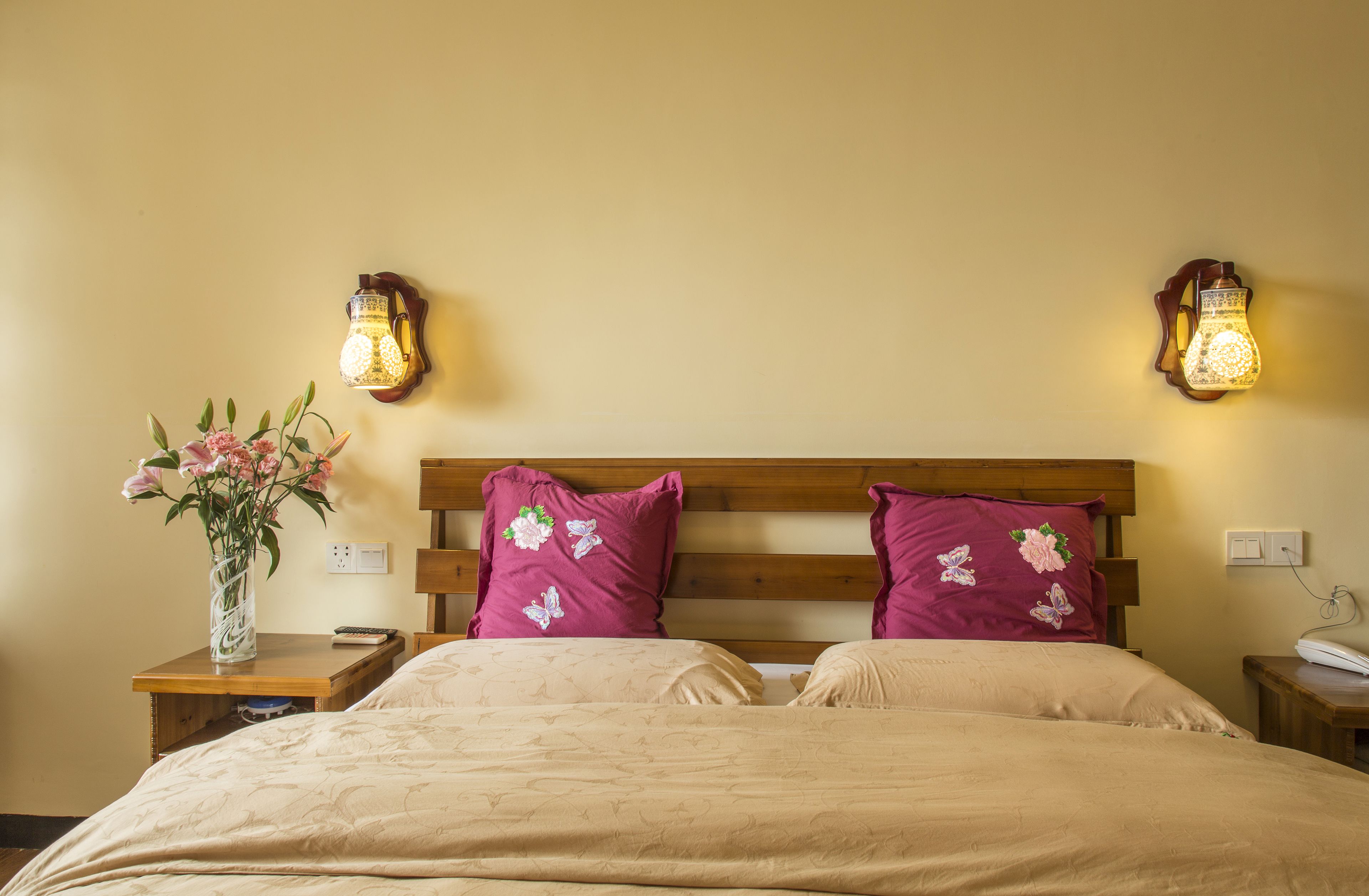 A hotel room with a wooden headboard, two wall-mounted lamps, a bedside table with a vase of flowers, and a bed with floral pillows.