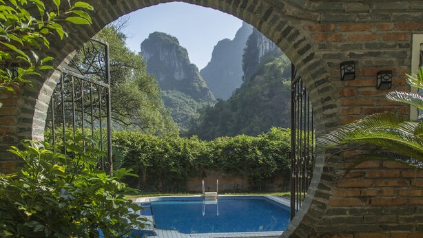 Una estructura de piedra arqueada que conduce a una piscina rodeada de exuberante vegetación y montañas.