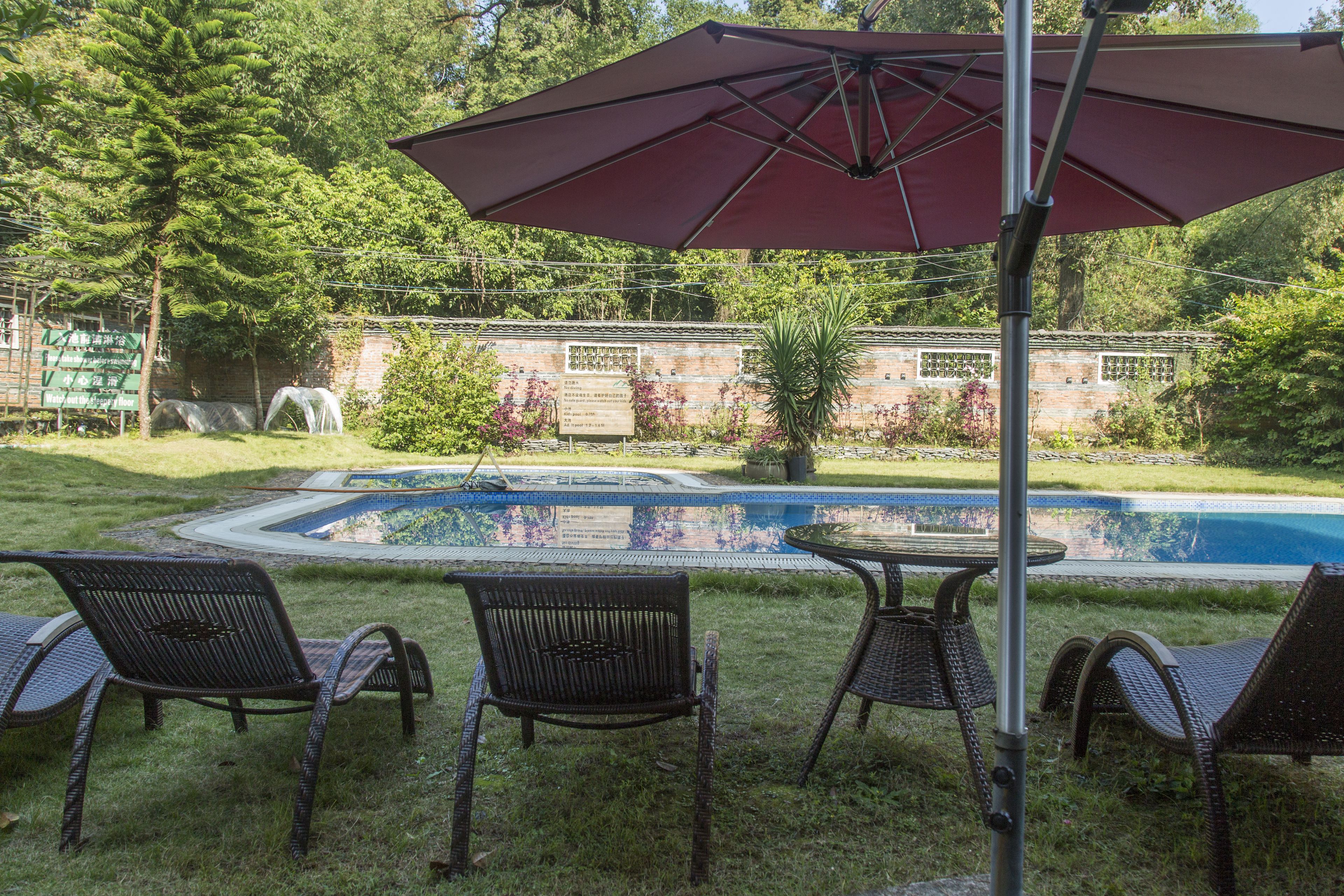A backyard with a swimming pool, surrounded by grass and trees. There are wicker chairs and a table with a red umbrella.