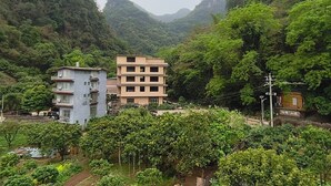 Un edificio de varios pisos con techo plano, rodeado de abundante vegetación y montañas al fondo.