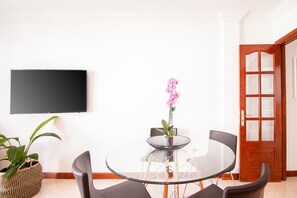 Ein Zimmer mit einem runden Glastisch, vier Stühlen, einem Blumentopf, einem Flachbildfernseher und einer Holztür.
