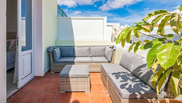A balcony with a wicker sofa, a small ottoman, and a potted plant.