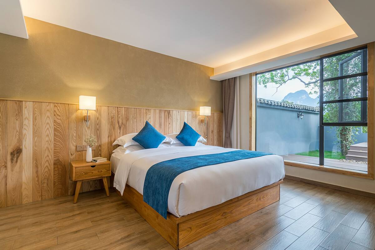 A modern bedroom with a wooden headboard, a large window with a view of greenery and mountains, a bedside table with a lamp, and a neatly made bed with blue bedding.
